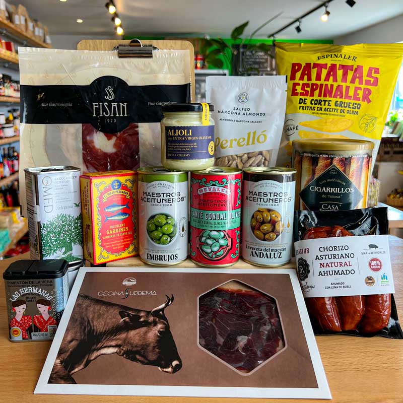 A curated gourmet food gift assortment displayed on a wooden counter inside a grocery store. The collection includes artisanal meats like Fisan Iberico Ham, Bienastur Smoked Chorizo Asturiano, and Entrepenas Cecina, canned olives, a jar of Alioli, and Patatas Espinaler chips.